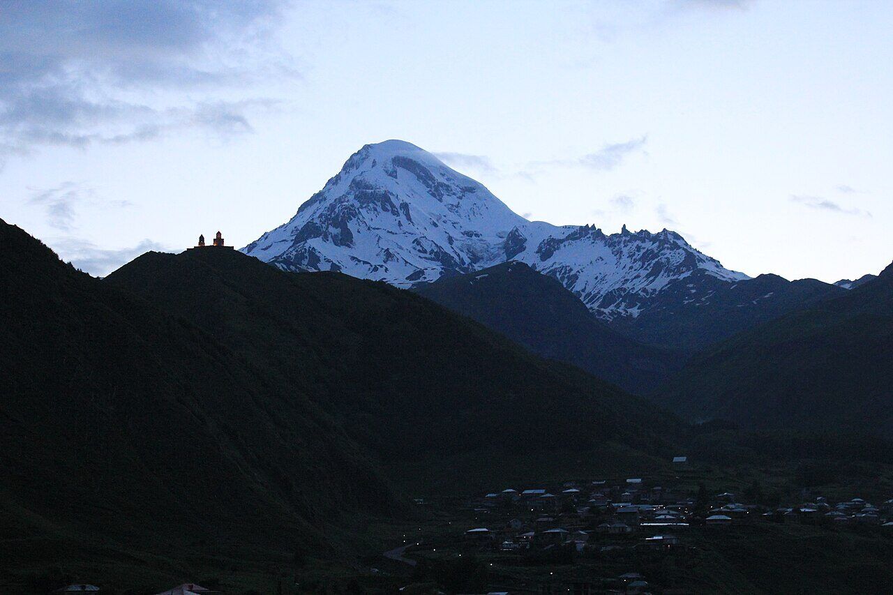 Gergeti Trinity Church on a ridge below the snow-capped peak of Mount Kazbek at dusk